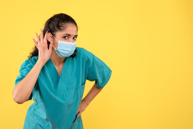 Vista frontale del medico abbastanza femminile con mascherina medica che ascolta qualcosa sulla parete gialla