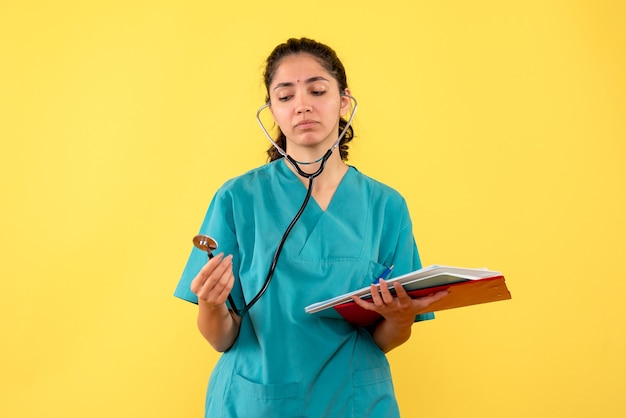 Vista frontale del medico della donna in uniforme che esamina i documenti della tenuta dello stetoscopio sulla parete gialla