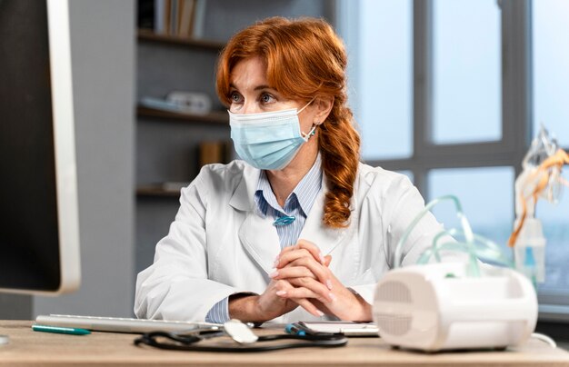 Vista frontale del medico femminile alla sua scrivania con mascherina medica guardando il computer