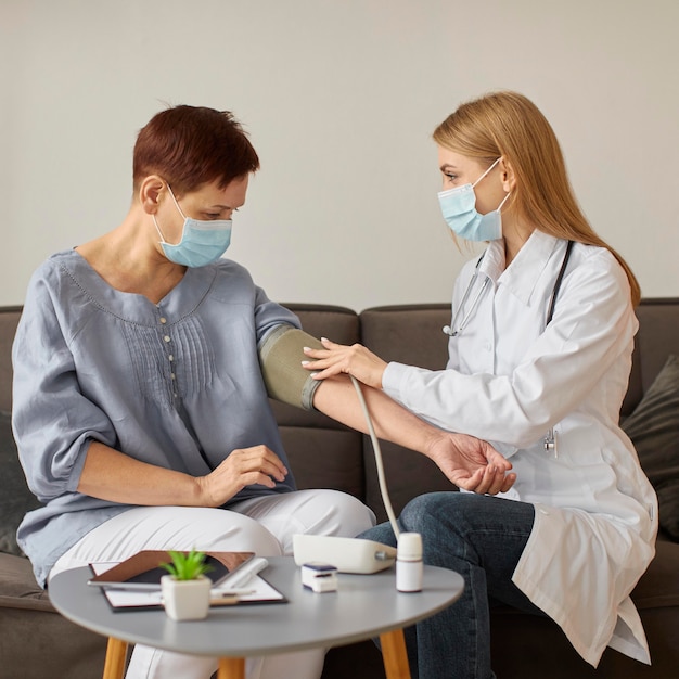 Vista frontale del medico femminile del centro di recupero covid con maschera medica che controlla la pressione sanguigna del paziente anziano