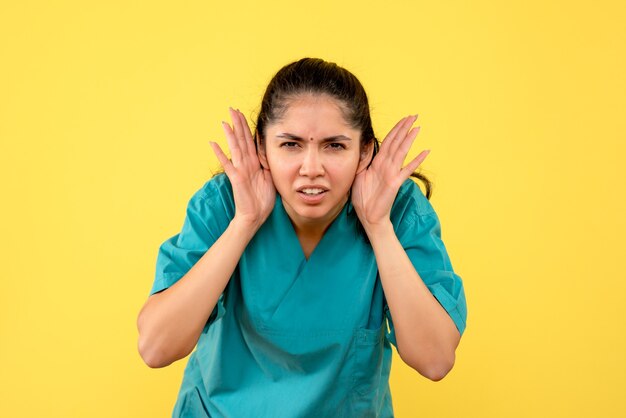 Vista frontale del medico femminile grazioso che mette le mani vicino alle sue orecchie sulla parete gialla