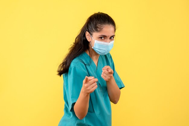 Vista frontale del medico femminile grazioso con la mascherina medica pointign nella parte anteriore sulla parete gialla