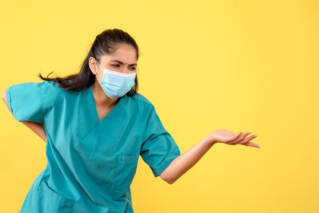 Vista frontale del medico femminile grazioso con mascherina medica che la tiene indietro sulla parete gialla