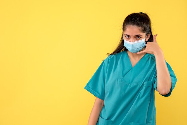 Vista frontale del medico femminile grazioso con mascherina medica che mi chiama segno sulla parete gialla