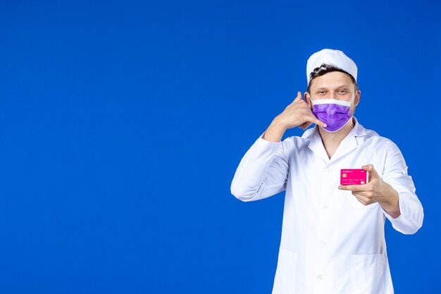 Vista frontale del medico maschio felice che tiene la carta di credito rosa imitando la telefonata sulla parete blu
