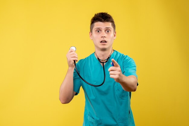 Vista frontale del medico maschio in tuta medica con lo stetoscopio sulla parete gialla