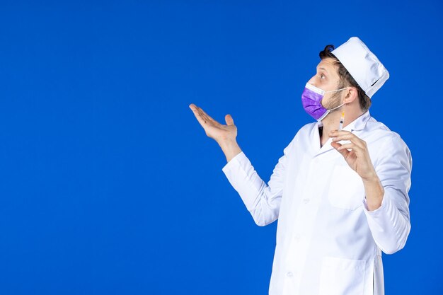 Vista frontale del medico maschio in tuta medica e maschera tenendo l'iniezione sull'azzurro