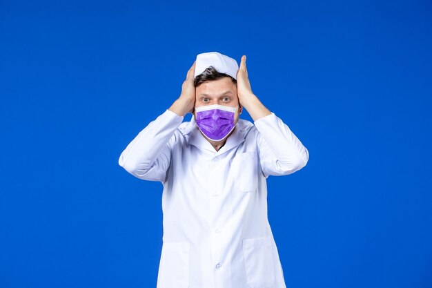 Vista frontale del medico maschio sollecitato in tuta medica e maschera viola sull'azzurro