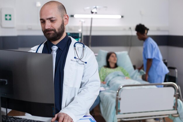 Vista frontale del medico professionista che digita competenza medica sul computer mentre è in background l'assistente nero che discute del trattamento sanitario. Paziente ricoverato con disturbi respiratori