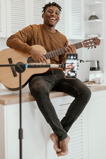 Vista frontale del musicista maschio di smiley a casa a suonare la chitarra e registrare con il telefono cellulare