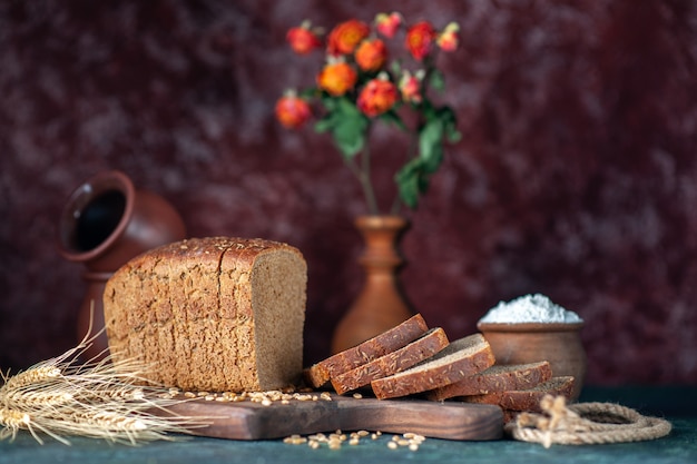 Vista frontale del pane nero dietetico picchi di grano su tagliere di legno ciotole vaso di fiori su sfondo blu marrone misti colori