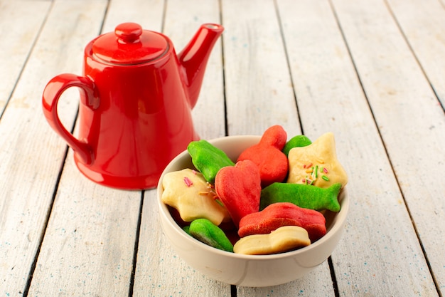 Vista frontale del piatto interno formato differente variopinto delizioso dei biscotti con il bollitore rosso sulla superficie grigia