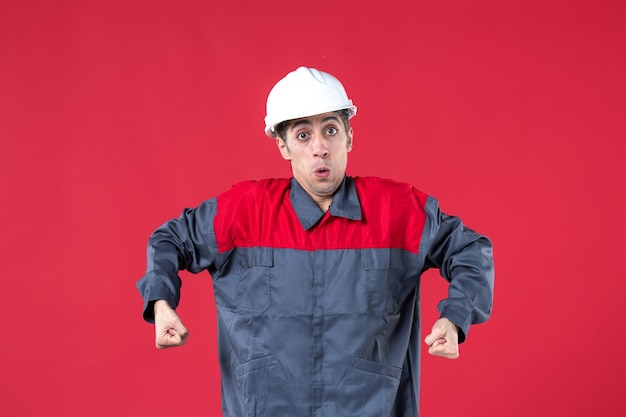 Vista frontale del preoccupante giovane lavoratore in uniforme con elmetto e stringendo i pugni sul muro rosso isolato