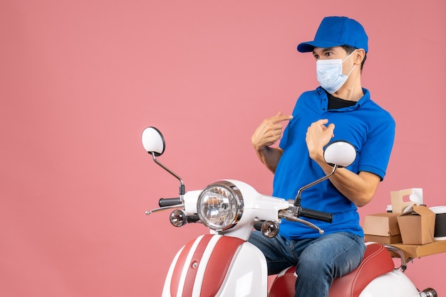 Vista frontale del ragazzo delle consegne confuso in maschera medica che indossa un cappello seduto su uno scooter su sfondo color pesca pastello