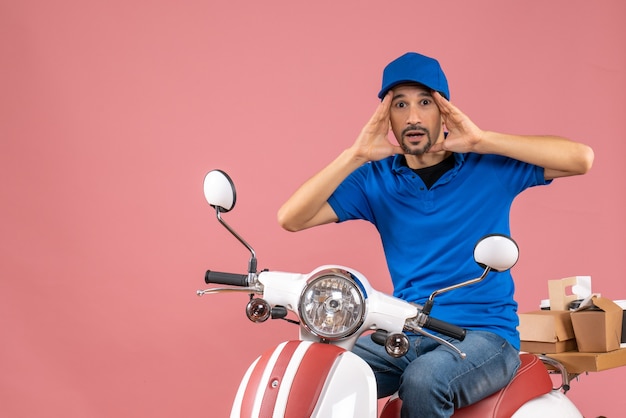 Vista frontale del ragazzo delle consegne sorpreso che indossa un cappello seduto su uno scooter su sfondo color pesca pastello