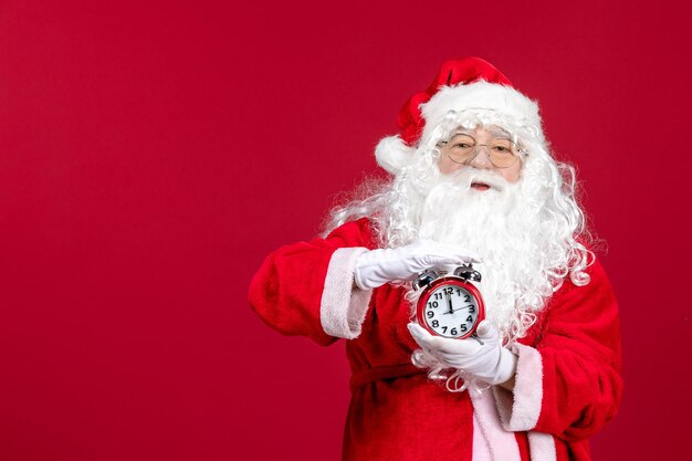 Vista frontale del vecchio babbo natale in abito rosso che tiene l'orologio sul muro rosso