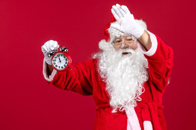 Vista frontale del vecchio babbo natale in abito rosso che tiene l'orologio sul muro rosso