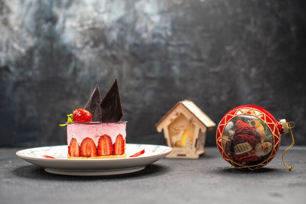Vista frontale deliziosa cheesecake con fragole e cioccolato su piastra ovale rossa lanterna a sfera albero di natale su buio