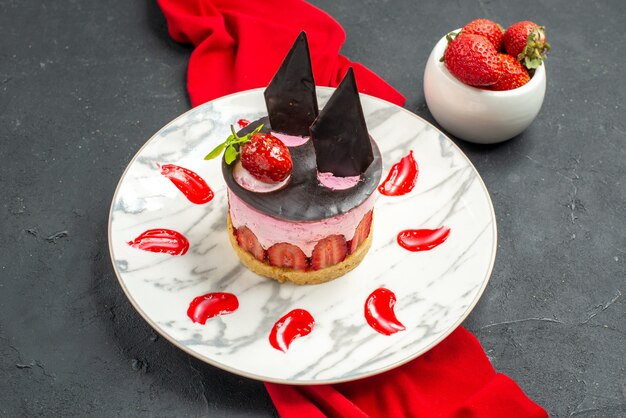 Vista frontale deliziosa cheesecake con fragole e cioccolato su piatto scialle rosso ciotola con fragole su oscurità