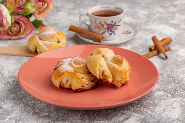 Vista frontale deliziosi pasticcini con ripieno all'interno della piastra insieme a tè e cannella sul tavolo bianco, torta cuocere alla frutta pasticceria