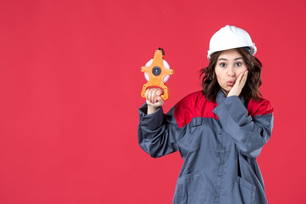 Vista frontale dell'architetto femminile confuso in uniforme con il nastro di misurazione della tenuta del cappello duro sulla parete rossa isolata