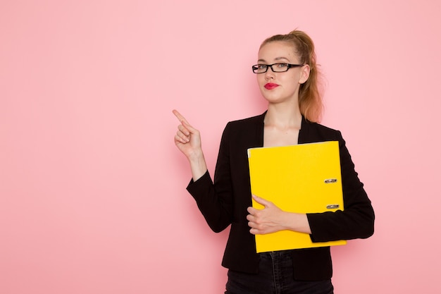 Vista frontale dell'impiegato femminile in giacca rigorosa nera che tiene documento giallo sulla parete rosa