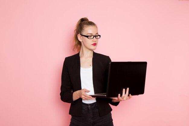 Vista frontale dell'impiegato femminile in giacca rigorosa nera che tiene e utilizza il suo computer portatile sulla parete rosa chiaro