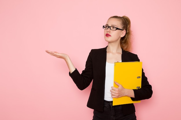 Vista frontale dell'impiegato femminile in giacca rigorosa nera che tiene file giallo sulla parete rosa chiaro