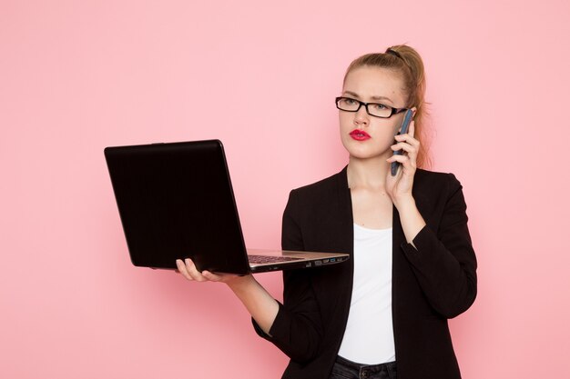 Vista frontale dell'impiegato femminile in giacca rigorosa nera che tiene utilizzando il computer portatile sulla parete rosa chiaro