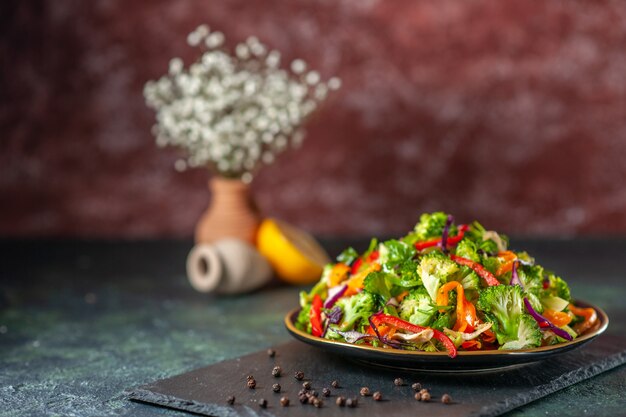 Vista frontale dell'insalata vegana con ingredienti freschi in un piatto e pepe su tagliere nero