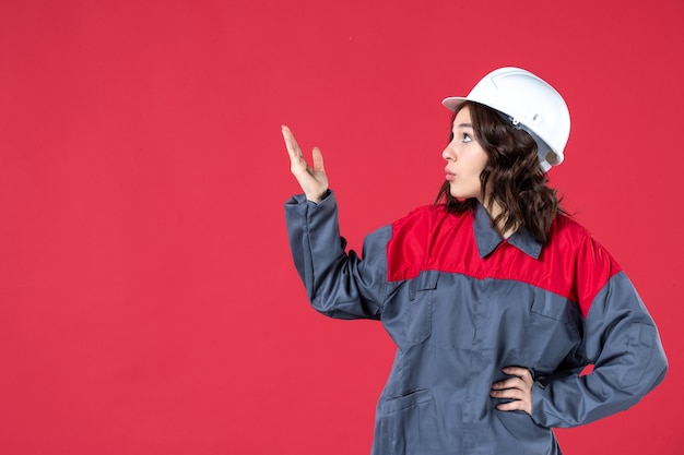 Vista frontale dell'interrogatorio del costruttore femminile in uniforme con elmetto e rivolto verso l'alto su sfondo rosso isolato