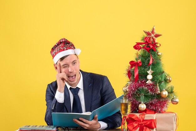 Vista frontale dell'occhio lampeggiante dell'uomo d'affari seduto al tavolo vicino all'albero di Natale e regali sulla parete gialla