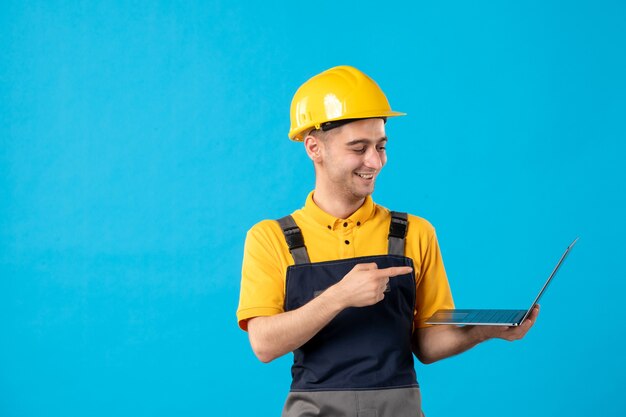 Vista frontale dell'operaio maschio di risata in uniforme con il computer portatile sulla superficie blu