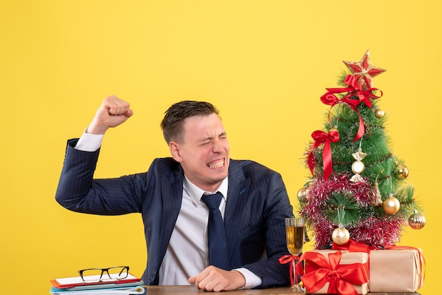 Vista frontale dell'uomo arrabbiato alzando la mano seduto al tavolo vicino all'albero di Natale e regali sulla parete gialla