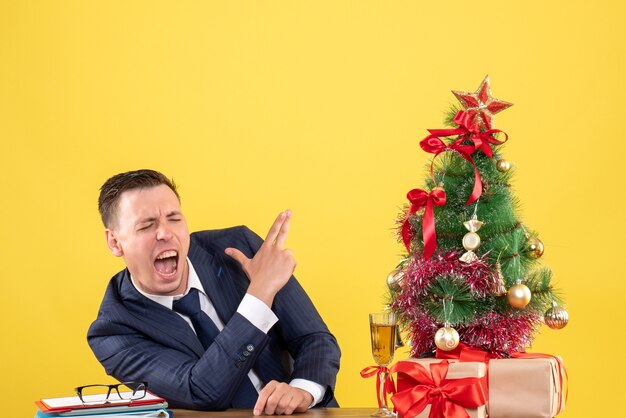 Vista frontale dell'uomo arrabbiato che fa il segno della pistola del dito che si siede al tavolo vicino all'albero di Natale e regali sulla parete gialla