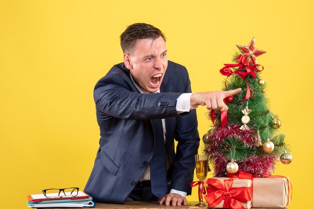 Vista frontale dell'uomo arrabbiato che grida a qualcuno in piedi dietro il tavolo vicino all'albero di Natale e regali sulla parete gialla
