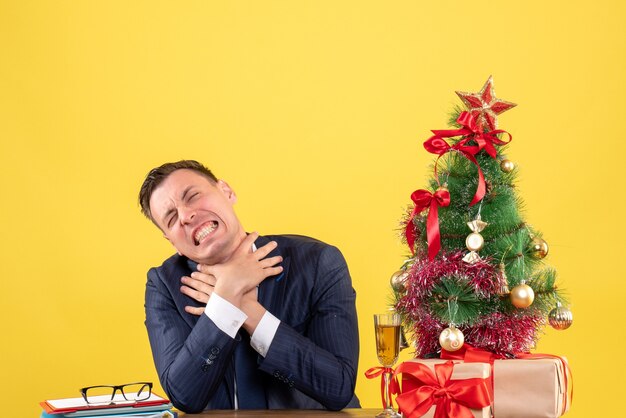 Vista frontale dell'uomo arrabbiato che si strangola con entrambe le mani seduto al tavolo vicino all'albero di Natale e regali sulla parete gialla
