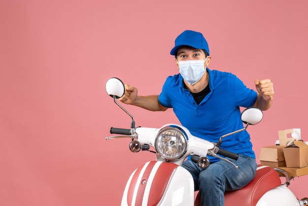 Vista frontale dell'uomo arrabbiato corriere nervoso in maschera medica che indossa cappello seduto su scooter su sfondo pesca pastello