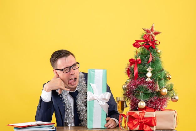 Vista frontale dell'uomo bello che fa mi chiama il segno del telefono che si siede al tavolo vicino all'albero di Natale e presenta su giallo