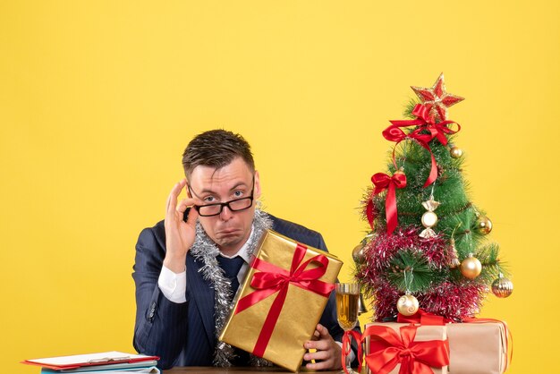 Vista frontale dell'uomo bello con gli occhiali che si siede al tavolo vicino all'albero di Natale e regali su giallo