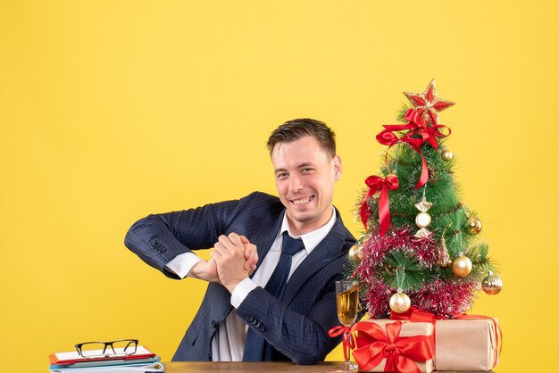 Vista frontale dell'uomo bello sorridente seduto al tavolo vicino all'albero di Natale e regali su giallo