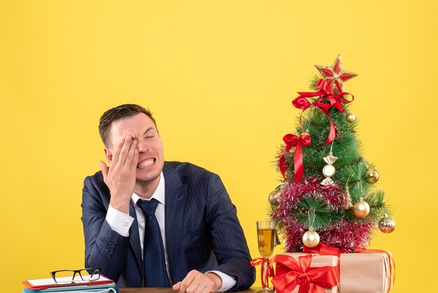 Vista frontale dell'uomo che piange che copre il suo occhio con la mano seduta al tavolo vicino all'albero di Natale e regali su giallo