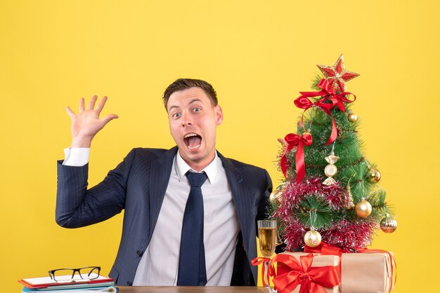 Vista frontale dell'uomo con gli occhi spalancati seduto al tavolo che dice ciao vicino all'albero di Natale e regali su giallo