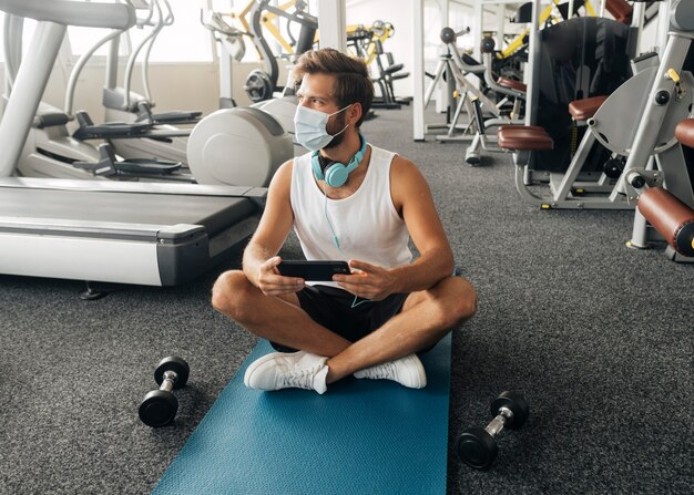 Vista frontale dell'uomo con le cuffie e la mascherina medica che tiene smartphone in palestra