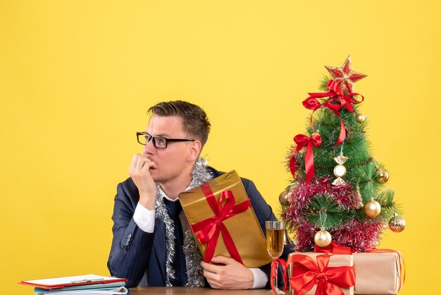 Vista frontale dell'uomo confuso guardando a sinistra seduto al tavolo vicino all'albero di Natale e presenta su giallo