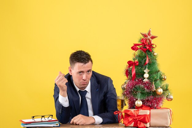 Vista frontale dell'uomo confuso seduto al tavolo vicino all'albero di Natale e regali su giallo