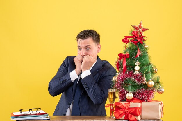 Vista frontale dell'uomo confuso seduto al tavolo vicino all'albero di Natale e regali su giallo
