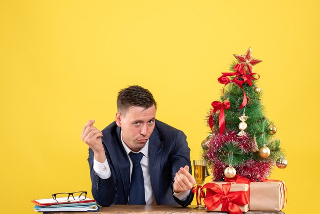 Vista frontale dell'uomo curioso che fa soldi segno seduto al tavolo vicino albero di Natale e regali su giallo