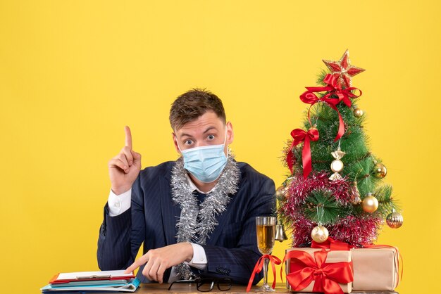 Vista frontale dell'uomo d'affari che punta in alto seduto al tavolo vicino all'albero di Natale e regali su giallo