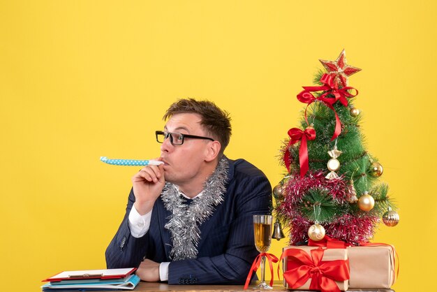 Vista frontale dell'uomo d'affari con gli occhiali utilizzando noisemaker vicino all'albero di Natale e regali su giallo